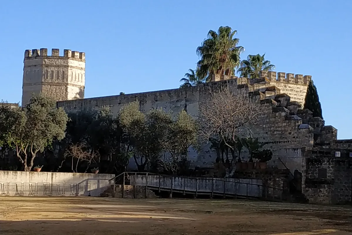 A crenellated stone wall and an octagonal tower of the Alcázar of Jerez rising above olive trees and a sandy courtyard