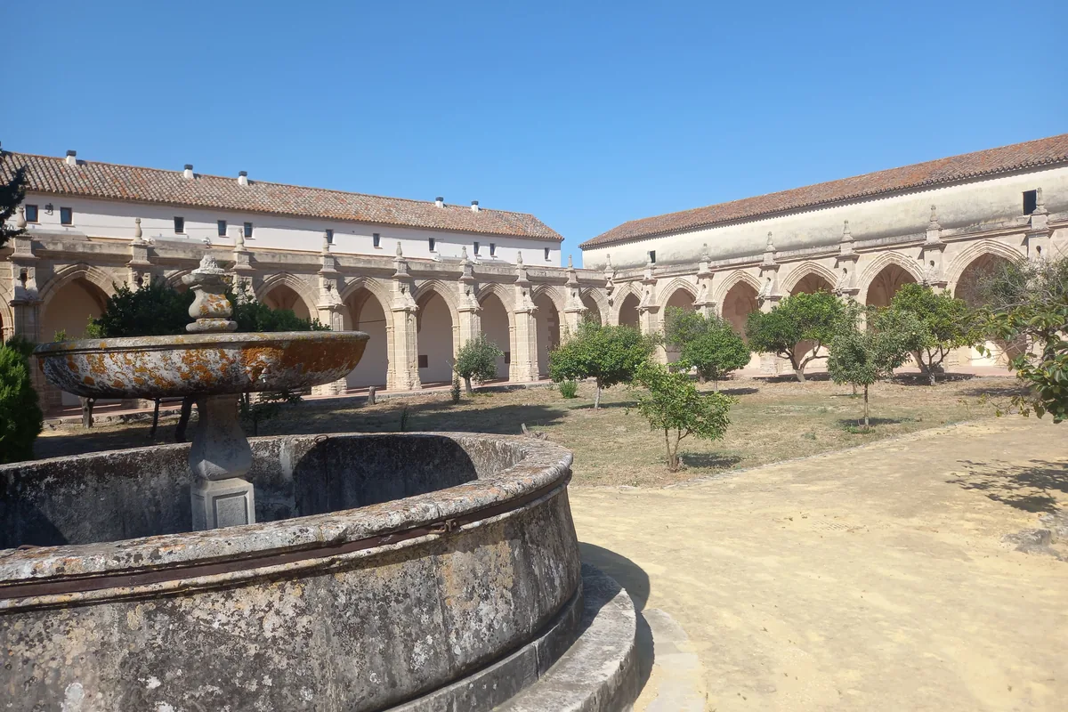 A long arcaded cloister of pale stone beside a circular stone basin, with orange trees on sandy ground
